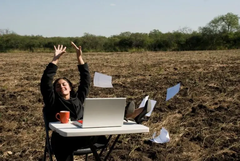 A woman sitting outdoors with her arms raised in joy, a laptop and coffee on the table, and papers flying in the wind, symbolizing relief and renewed energy after overcoming Candida overgrowth.