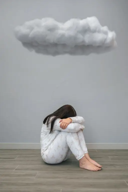 A woman sitting on the floor in a curled-up position with her head down, symbolizing depression and fatigue, with a dark cloud hovering above her, representing mental and emotional symptoms of Candida overgrowth.