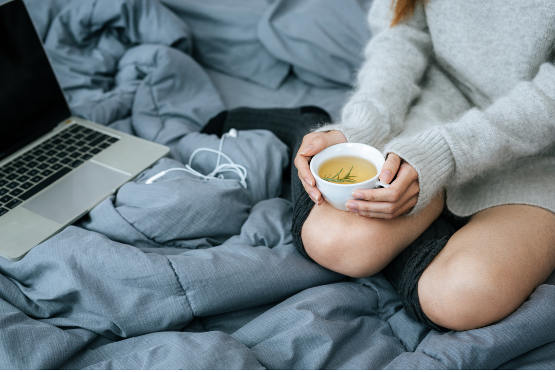 Individual relaxing on a cozy bed with a cup of herbal tea, laptop nearby to enjoy the their exclusive wellness benefits