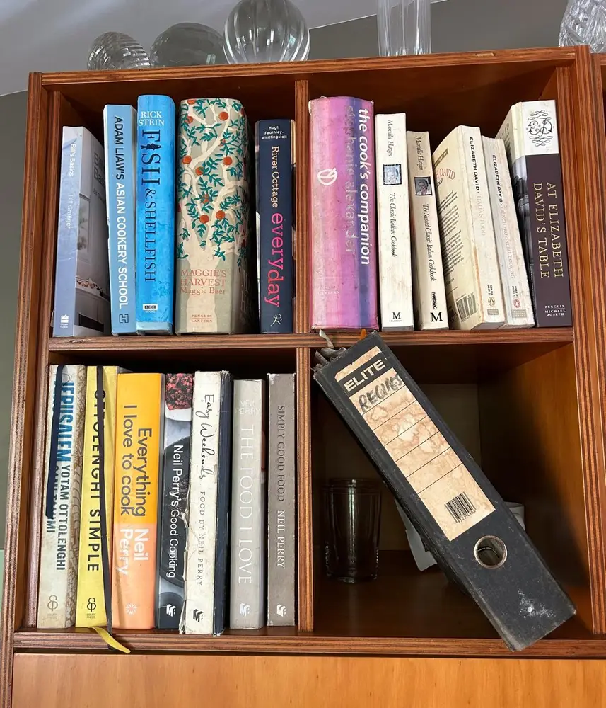 A wooden bookshelf filled with a variety of cookbooks, with a black binder labeled 'Recipes' in the bottom right corner.